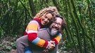 Ein Paar lächelt in die Kamera. Die Frau wird von ihrem Partner Huckepack durch den Wald getragen. | Bild: mauritius images / Fabio and Simona