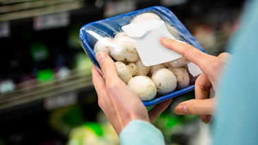 Frau hält in einem Supermarkt eine Packung mit Champignons in der Hand | Bild: mauritius images