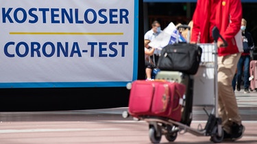 Mann schiebt am Flughafen München einen Trolley mit Koffern an dem Schild mit der Aufschrift "Kostenloser Coronatest" vorbei | Bild: dpa/picture alliance