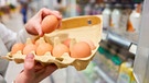 Eine Hand hält in einem Supermarkt einen Eierkarton mit zehn Eiern und überprüft, ob sie noch ganz sind. | Bild: mauritius-images