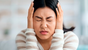 Eine Frau hält sich mit schmerzverzerrtem Gesicht die Ohren zu. | Bild: mauritius images / Prostock-studio / Alamy / Alamy Stock Photos