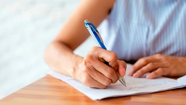 Eine Frau hält einen blauen Kugelschreiber in einer Hand und notiert etwas auf einem weißen Blatt. | Bild: mauritius images / Kittipong Satrinekarn / Alamy / Alamy Stock Photos