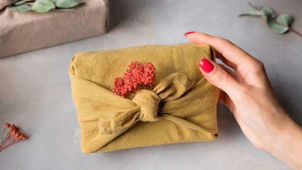 Eine weibliche Hand mit rot lackierten Fingernägeln hält ein Geschenk, dass in einem Tuch verpackt ist | Bild: mauritius-images