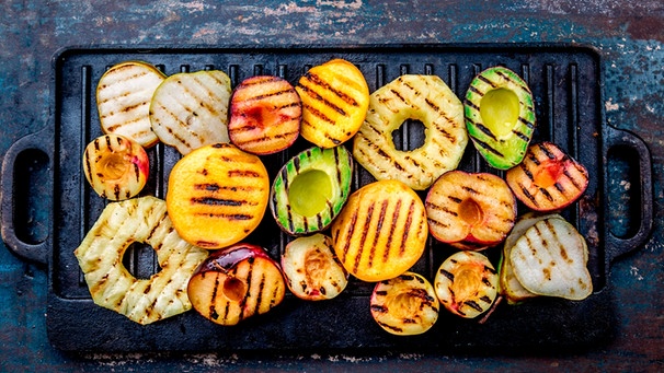 Auf einem Grill liegen gegrillte Avocadohälften und Obst. | Bild: mauritius-images