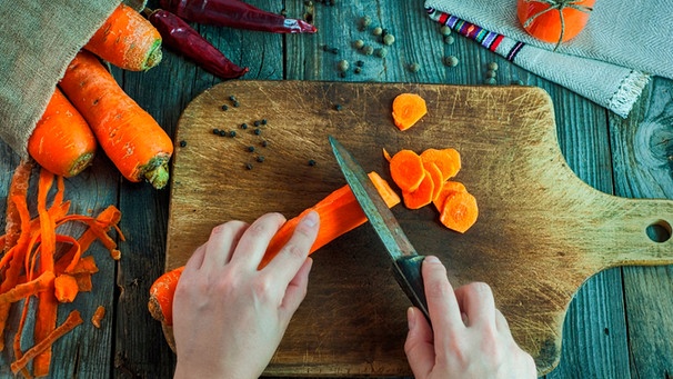 Karotten mit grünen Köpfen liegen neben einem Schneidebrett, eine Hand schneidet Karotten in Scheiben | Bild: mauritius images