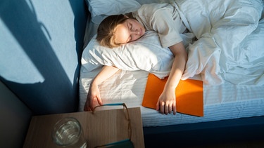 Frau liegt im Bett und schläft, neben sich ein Buch | Bild: mauritius images