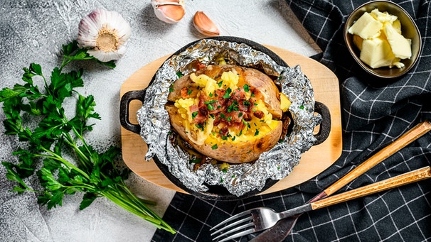 Auf einer gusseisernen Pfanne liegt eine aus Alufolie ausgepackte Ofenkartoffel mit Speckwürfeln. Um die Pfanne liegt ein Bund Petersilie, eine Konblauchknolle, Butter und Besteck. | Bild: mauritius-images