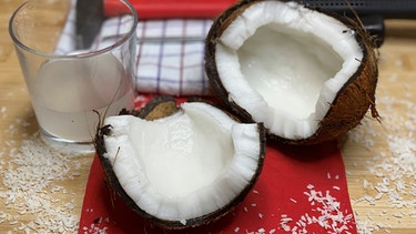 Zwei Hälften einer geöffneten Kokosnuss liegen neben einem Glas mit Kokoswasser auf einem Tisch. | Bild: Jonas Schramm, BR