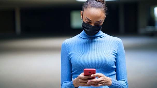 Eine junge Frau blickt auf ihr Handy. Sie trägt eine Maske. | Bild: mauritius-images