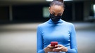 Eine junge Frau blickt auf ihr Handy. Sie trägt eine Maske. | Bild: mauritius-images
