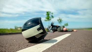 Symbolbild Motorradunfall: Motorradhelm liegt auf einer Straße, im Hinergrund liegt ein verunfallter Motorradfahrer | Bild: mauritius images / Joerg Huettenhoelscher / Alamy / Alamy Stock Photos
