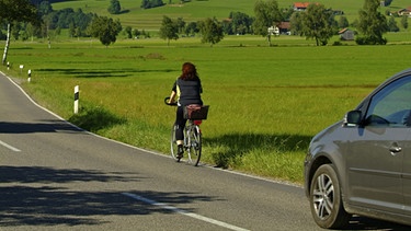 Pkw fährt hinter einer Radfahrerin auf einer Landstraße her | Bild: dpa/picture alliance
