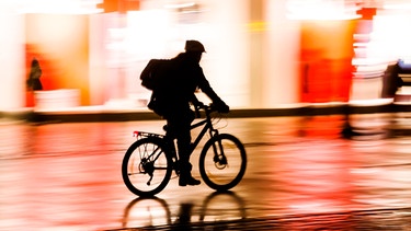Radfahrer fährt nachts in der Stadt an beleuchteten Schaufenstern vorbei | Bild: mauritius images