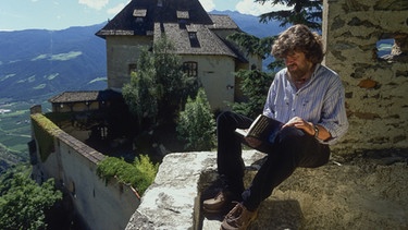 Reinhold Messner 2001 sitzt mit einem Buch auf der Mauer von Schloss Juval, seinem Sommerwohnsitz. | Bild: dpa/picture alliance