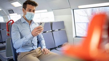 Mann sitzt mit Maske und Smartphone sitzt in einem Zug | Bild: mauritius images