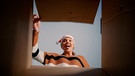 Eine Frau öffnet einen Karton und blickt strahlend hinein. | Bild: mauritius images / Dragos Condrea / Alamy / Alamy Stock Photos