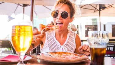 Eine Frau beißt in ein Stück Pizza vor ihr stehen ein Glas mit Bier und eins mit Cola. | Bild: mauritius-images