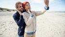 Paar steht am Strand und grüßt ins Handy bei einem Videoanruf | Bild: mauritius images