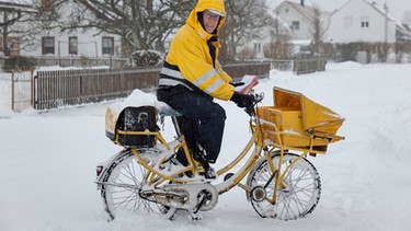 Postzusteller steht mit seinem Fahrrad im Schnee. | Bild: mauritius images