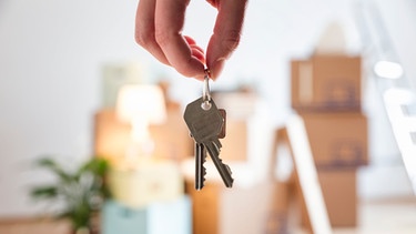Hand einer Frau hält Wohnungsschlüssel, im Hintergrund stehen Umzugskartons. | Bild: mauritius images