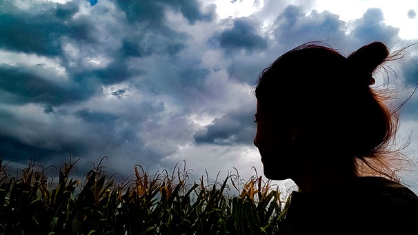 Eine Frau steht vor einem Maisfeld und schaut in den Himmel voller Gewitterwolken | Bild: mauritius-images