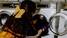Eine Frau gibt ihre Wäsche in die Waschmaschine, dabei trägt sie ein Ballkleid. | Bild: Picaro Photography