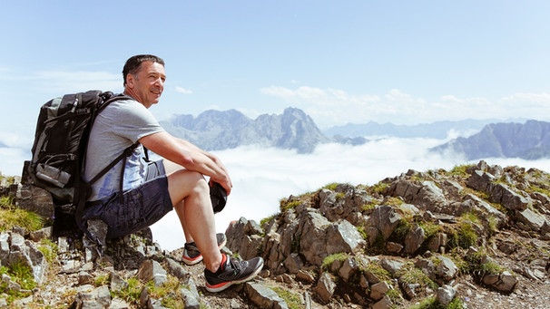 Ein Mann Mitte 50 sitzt auf einem Berggipfel und denkt nach | Bild: mauritius images