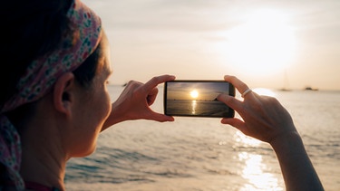 Frau fotografiert einen Sonnenuntergang am Meer im Urlaub | Bild: mauritius images