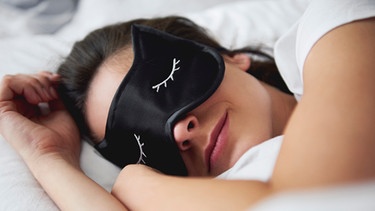 Frau liegt in einem Bett und schläft mit einer Schlafmaske | Bild: mauritius images