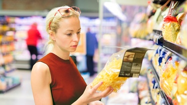 Frau liest sich in einem Supermarkt die Zutatenliste auf einer Nudelpackung durch | Bild: mauritius images