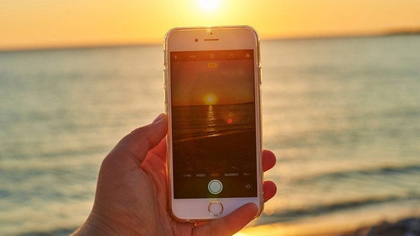 Mann fototgrafiert Sonnenuntergang am Strand mit dem Smartphone | Bild: mauritius images