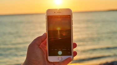 Mann fototgrafiert Sonnenuntergang am Strand mit dem Smartphone | Bild: mauritius images