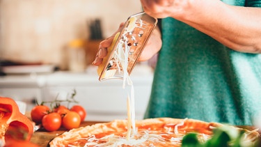 Frau in einer Küche reibt Käse auf eine Pizza  | Bild: mauritius images Zivica Kerkez