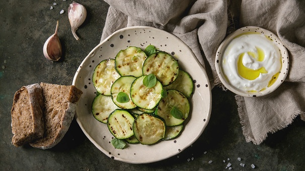 Ein Teller mit einem Salat aus gerösteten Zucchinischeiben steht mit einem Dip auf einem Holztisch | Bild: mauritius images