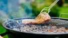 Ein Stück Fleisch wird auf einem heißen Grill mit einer Grillzange gewendet. | Bild: mauritius images / Westend61 / Pro Pix
