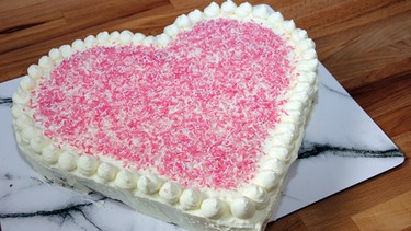 Ein Herzkuchen ist mit rosafarbenen Zuckerstreuseln und Sahne verziert.  | Bild: BR