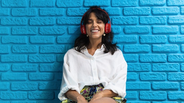 Eine junge Frau hört, an einer blauen Wand lehend, Musik über rote Kopfhörer. | Bild: mauritius-images