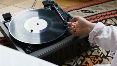 Auf einem Plattenspieler liegt eine Schallplatte. Eine Hand platziert den Tonarm auf der Platte. | Bild: mauritius-images