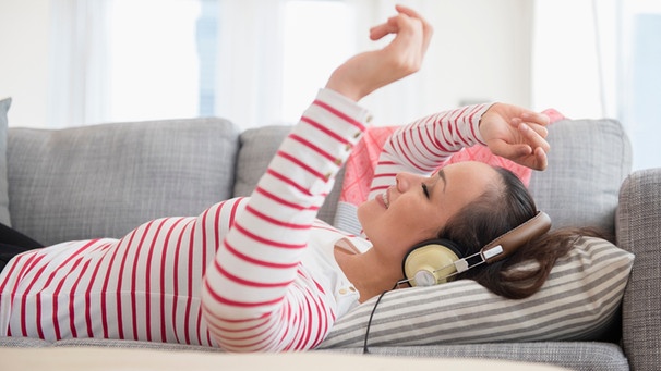 Frau liegt mit Kopfhörern auf der Couch und hört Musik übers Handy | Bild: mauritius-images