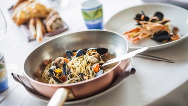 Eine Kupferpfanne steht auf dem Tisch. In der Pfanne und auf einem Teller liegen Spaghetti mit Muscheln.  | Bild: mauritius-images