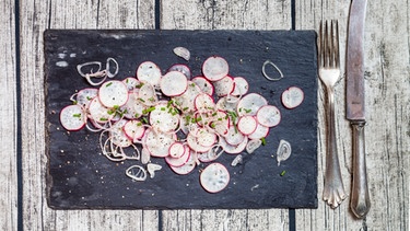 Auf einer Schieferplatte ist ein Radieschensalat mit Zwiebeln angerichtet | Bild: mauritius-images