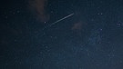 Eine Sternschnuppe fliegt über den dunklen Nachthimmel. | Bild: picture-alliance/dpa
