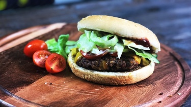 Ein vegetarischer Burger mit Käse, Salat und Tomate liegt auf einem Holzbrett. | Bild: BR