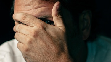 Mann versteckt Gesicht hinter seiner Hand und schaut vorsichtig durch zwei Finger der Hand. | Bild: mauritius images  Loop Images  Mickey Strider