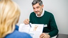 Ein plastischer Chirurg erläutert einer Frau anhand eines Fachbuches den geplanten Eingriff an der Brust. | Bild: picture-alliance/dpa