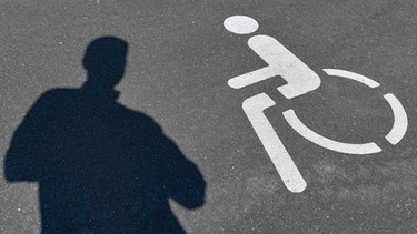 Ein Piktogramm für einen Behindertenparkplatz und ein Schatten einer Person sind auf dem Asphalt zu sehen.  | Bild: picture-alliance/dpa