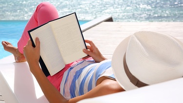 Eine Frau liest ein Buch, sie trägt einen Sonnenhut, im Hintergrund das Meer | Bild: colourbox.com