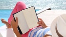 Eine Frau liest ein Buch, sie trägt einen Sonnenhut, im Hintergrund das Meer | Bild: colourbox.com