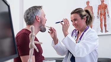 Eine Hausärztin schaut ihrem Patienten mithilfe eines Zungenspatels in den Mund. | Bild: picture-alliance/dpa