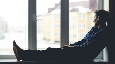 Ein Mann sitzt auf dem Fensterbrett und sieht aus dem Fenster. | Bild: stock.adobe.com/Mihail